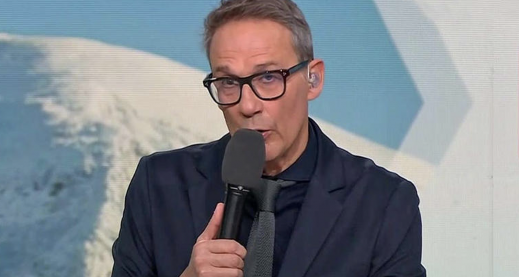 Animateur de télévision en studio tenant un micro noir près de la bouche, portant des lunettes à monture noire, une veste bleu marine et une cravate sombre, oreillette transparente à l’oreille droite, expression concentrée et regard appuyé face caméra, décor de plateau aux tons bleus avec fond graphique flouté en arrière-plan.