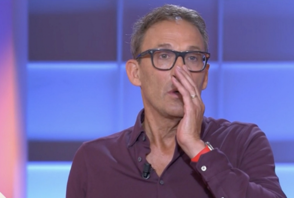 Animateur de télévision en plein direct sur un plateau lumineux aux tons bleus et violets, portant des lunettes à monture noire et une chemise bordeaux légèrement ouverte au col, micro-cravate fixé sur la poitrine, la main droite posée contre sa bouche dans un geste de surprise ou de tension, alliance visible au doigt et bracelet rouge au poignet, expression sérieuse et concentrée, décor de studio flouté en arrière-plan avec panneaux lumineux rectangulaires.