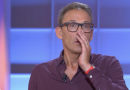 Animateur de télévision en plein direct sur un plateau lumineux aux tons bleus et violets, portant des lunettes à monture noire et une chemise bordeaux légèrement ouverte au col, micro-cravate fixé sur la poitrine, la main droite posée contre sa bouche dans un geste de surprise ou de tension, alliance visible au doigt et bracelet rouge au poignet, expression sérieuse et concentrée, décor de studio flouté en arrière-plan avec panneaux lumineux rectangulaires.