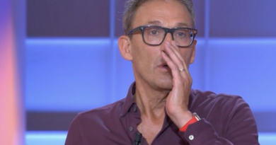 Animateur de télévision en plein direct sur un plateau lumineux aux tons bleus et violets, portant des lunettes à monture noire et une chemise bordeaux légèrement ouverte au col, micro-cravate fixé sur la poitrine, la main droite posée contre sa bouche dans un geste de surprise ou de tension, alliance visible au doigt et bracelet rouge au poignet, expression sérieuse et concentrée, décor de studio flouté en arrière-plan avec panneaux lumineux rectangulaires.