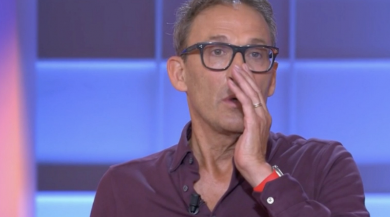 Animateur de télévision en plein direct sur un plateau lumineux aux tons bleus et violets, portant des lunettes à monture noire et une chemise bordeaux légèrement ouverte au col, micro-cravate fixé sur la poitrine, la main droite posée contre sa bouche dans un geste de surprise ou de tension, alliance visible au doigt et bracelet rouge au poignet, expression sérieuse et concentrée, décor de studio flouté en arrière-plan avec panneaux lumineux rectangulaires.
