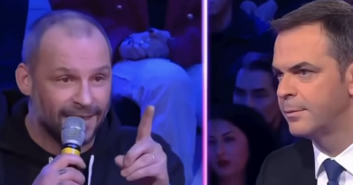 Un restaurateur chauve, tenant un micro à la main et pointant son doigt avec fermeté, s’exprime avec intensité sur un plateau télévisé face à un homme en costume sombre qui l’écoute attentivement. L’ambiance est tendue, avec un public en arrière-plan sous éclairage bleu et violet typique d’un talk-show politique.