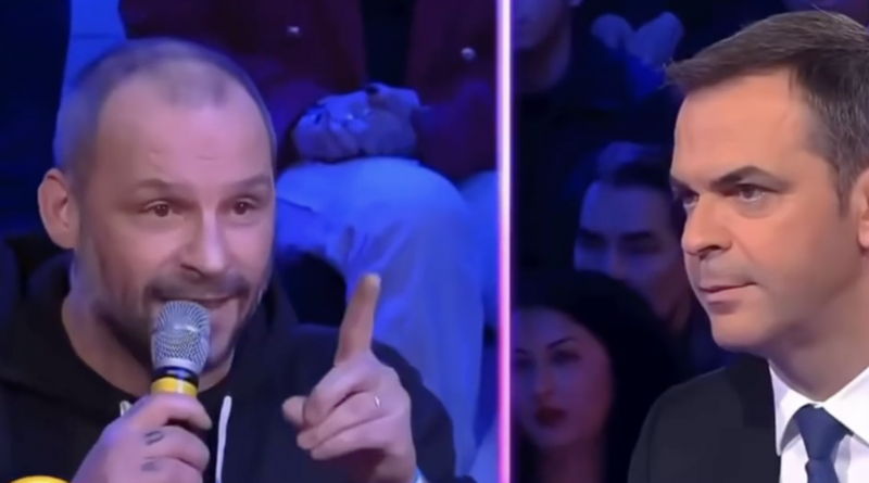 Un restaurateur chauve, tenant un micro à la main et pointant son doigt avec fermeté, s’exprime avec intensité sur un plateau télévisé face à un homme en costume sombre qui l’écoute attentivement. L’ambiance est tendue, avec un public en arrière-plan sous éclairage bleu et violet typique d’un talk-show politique.