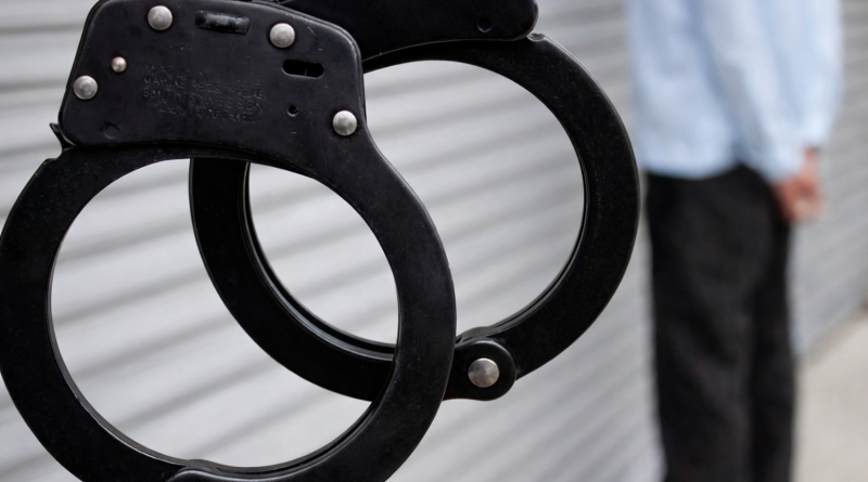 Photographie en gros plan d’une paire de menottes noires en métal, suspendues et légèrement ouvertes, nettes au premier plan, tandis qu’une silhouette masculine floue se tient en arrière-plan devant un mur clair à rainures horizontales, suggérant une scène liée à une interpellation ou à une procédure judiciaire.