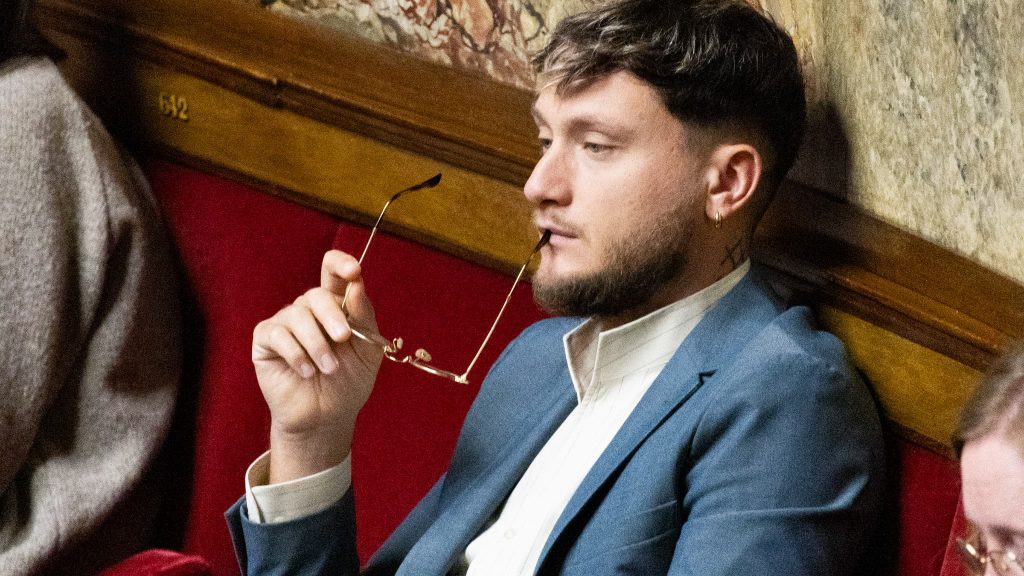 Jeune député assis sur un banc rouge de l’Assemblée nationale, costume bleu et chemise claire, tenant une paire de lunettes à la main dans une posture réfléchie.