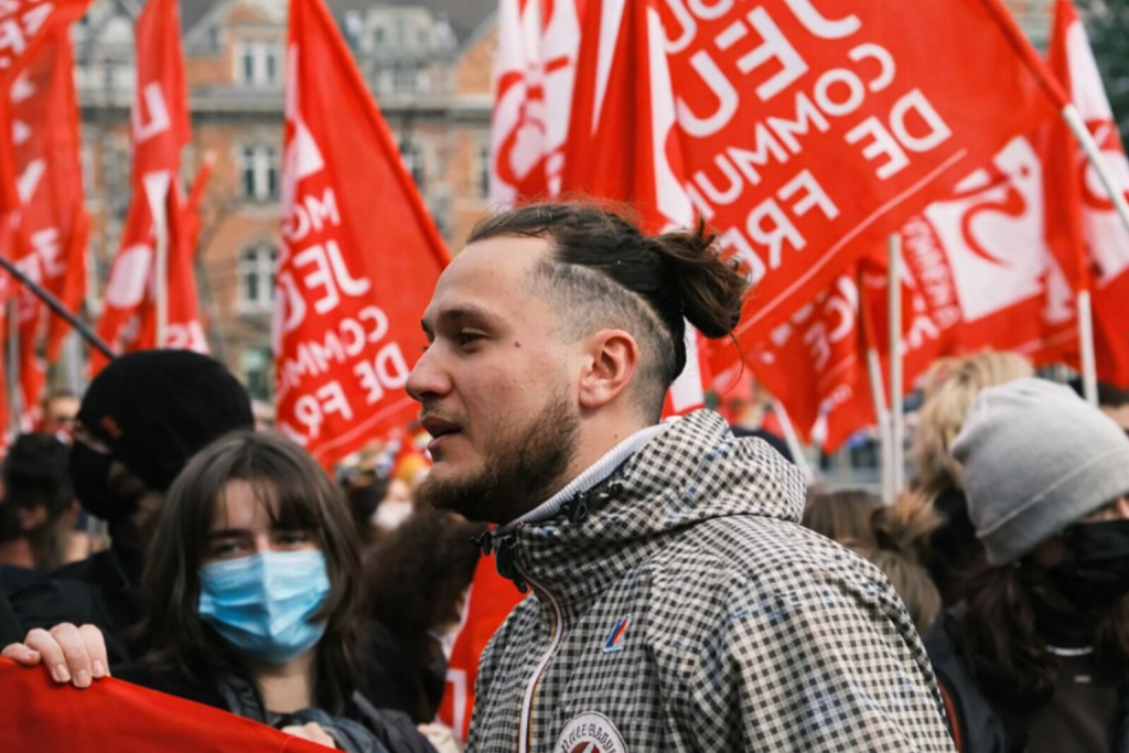 Un jeune homme aux cheveux attachés en chignon et à la barbe courte s’exprime lors d’une manifestation en plein air, entouré d’une foule de participants brandissant de nombreux drapeaux rouges avec des inscriptions blanches. Il porte une veste à motifs et regarde vers la droite avec un air déterminé. Autour de lui, plusieurs personnes sont visibles, certaines portant un masque chirurgical ou un bonnet. À l’arrière-plan, des bâtiments urbains anciens sont légèrement floutés, soulignant l’ambiance d’un rassemblement politique ou syndical en centre-ville.