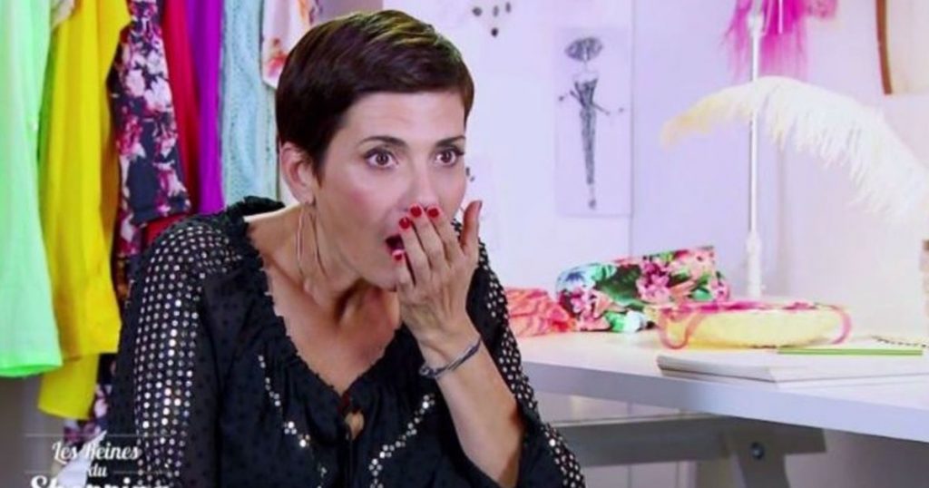 Animatrice de télévision assise dans une pièce colorée, la main devant la bouche, exprimant une vive surprise face à une situation inattendue.