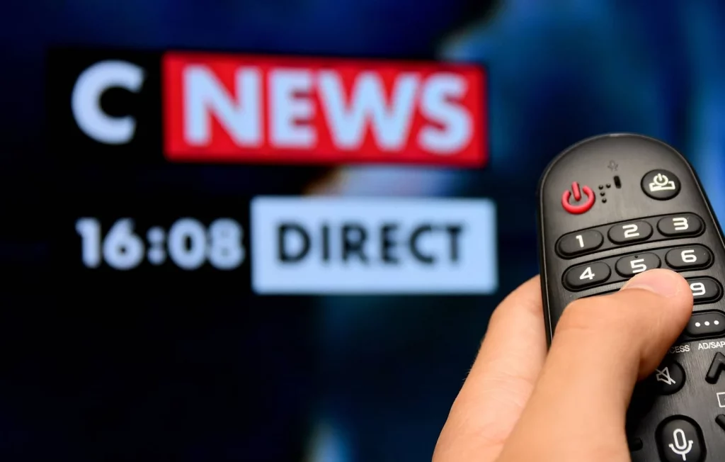 Télécommande tenue à la main face à un écran de télévision diffusant une chaîne d’information en direct, image volontairement floutée.