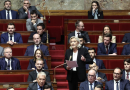 Une responsable politique prend la parole à la tribune au centre de l’hémicycle, tenant des feuilles en main, tandis que de nombreux députés, hommes et femmes en costume, sont assis sur les bancs rouges de l’Assemblée nationale lors d’un débat parlementaire.