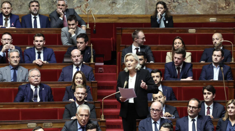 Une responsable politique prend la parole à la tribune au centre de l’hémicycle, tenant des feuilles en main, tandis que de nombreux députés, hommes et femmes en costume, sont assis sur les bancs rouges de l’Assemblée nationale lors d’un débat parlementaire.