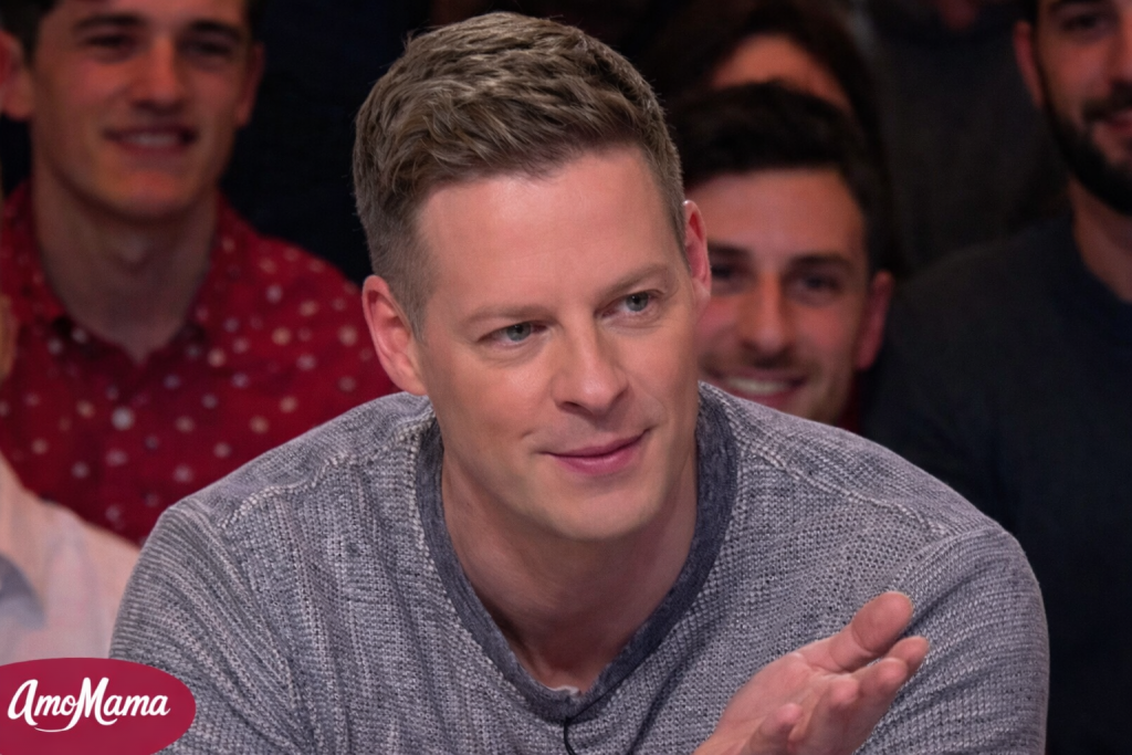 Chroniqueur de télévision assis sur un plateau, expression attentive, main levée en geste explicatif, public flouté en arrière-plan