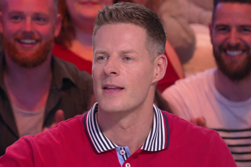 Chroniqueur de télévision assis sur un plateau, regard concentré, public souriant en arrière-plan, ambiance d’émission en direct