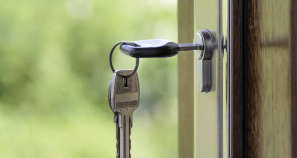 Clé métallique insérée dans la serrure d’une porte vitrée, trousseau suspendu, arrière-plan flou verdoyant suggérant un extérieur lumineux