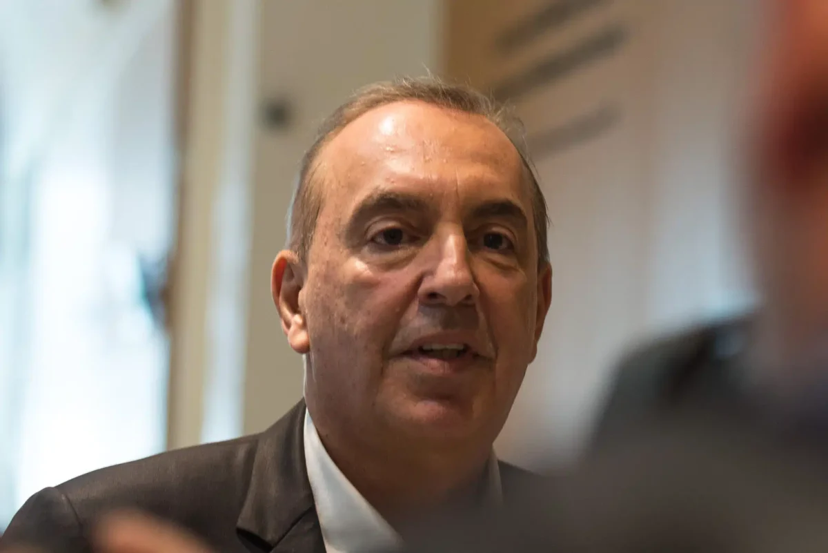 Portrait serré d’un homme mûr en costume sombre, assis à l’intérieur, expression grave, regard tourné vers son interlocuteur, arrière-plan flou évoquant un environnement professionnel.