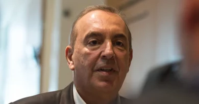 Portrait serré d’un homme mûr en costume sombre, assis à l’intérieur, expression grave, regard tourné vers son interlocuteur, arrière-plan flou évoquant un environnement professionnel.