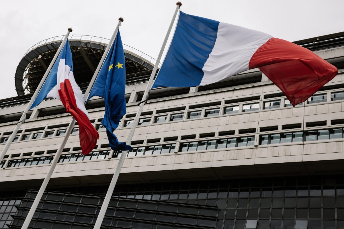 Drapeaux tricolores français et drapeau européen flottant devant un bâtiment administratif moderne en pierre et verre sous un ciel gris, symbolisant les institutions publiques et la gestion des finances nationales.