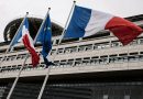 Drapeaux tricolores français et drapeau européen flottant devant un bâtiment administratif moderne en pierre et verre sous un ciel gris, symbolisant les institutions publiques et la gestion des finances nationales.