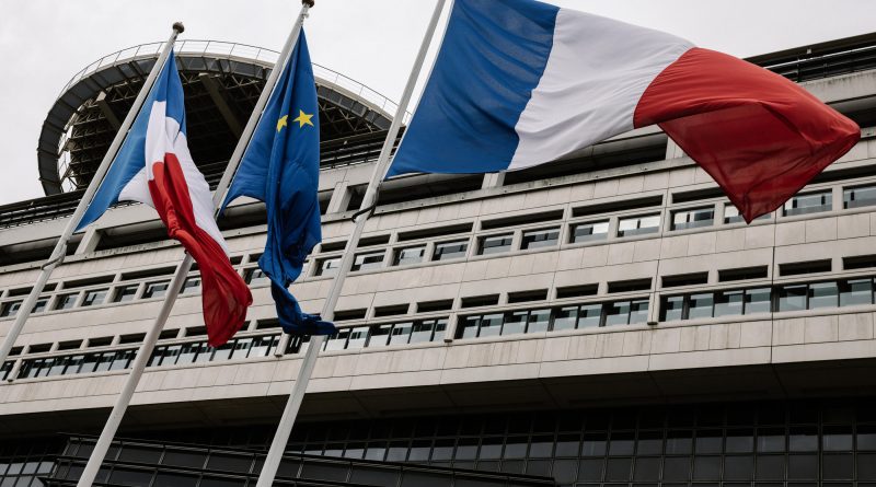 Drapeaux tricolores français et drapeau européen flottant devant un bâtiment administratif moderne en pierre et verre sous un ciel gris, symbolisant les institutions publiques et la gestion des finances nationales.