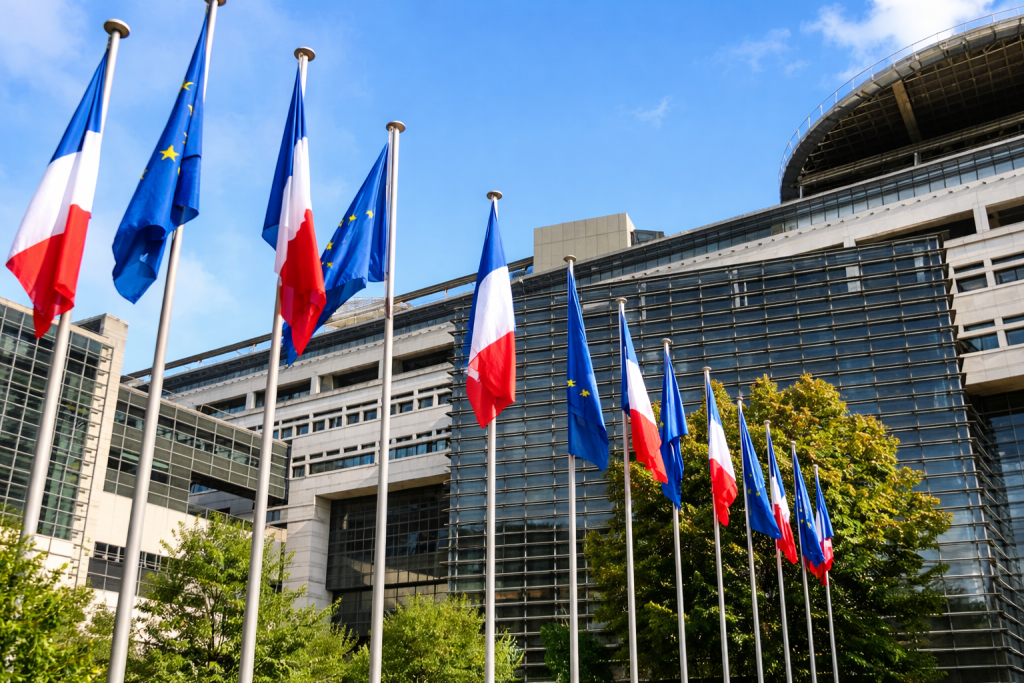 Drapeaux français et européens alignés devant un bâtiment institutionnel moderne en verre et en acier sous un ciel bleu, symbolisant les institutions publiques et la gouvernance nationale.