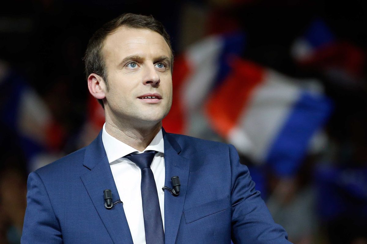 Un homme politique en costume bleu, debout derrière un pupitre, regard tourné vers l’assistance, avec des drapeaux tricolores flous en arrière-plan lors d’un discours public