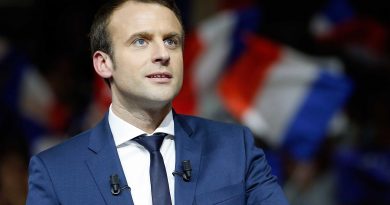 Un homme politique en costume bleu, debout derrière un pupitre, regard tourné vers l’assistance, avec des drapeaux tricolores flous en arrière-plan lors d’un discours public