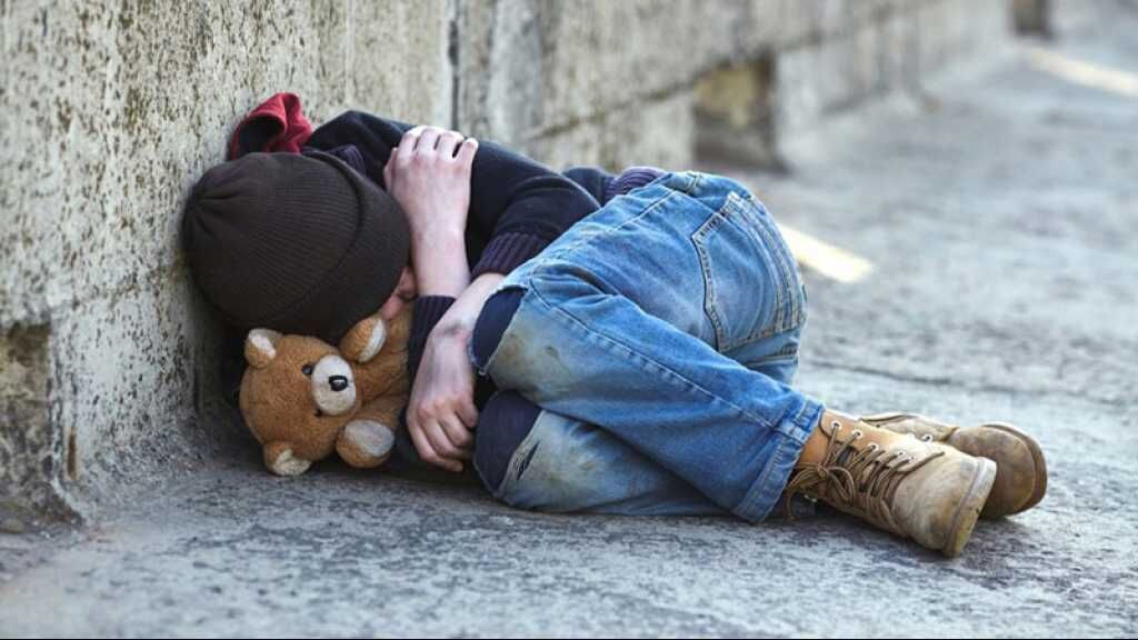 Enfant allongé sur le sol contre un mur, recroquevillé, tenant une peluche, vêtu de vêtements usés, dans un environnement urbain froid.