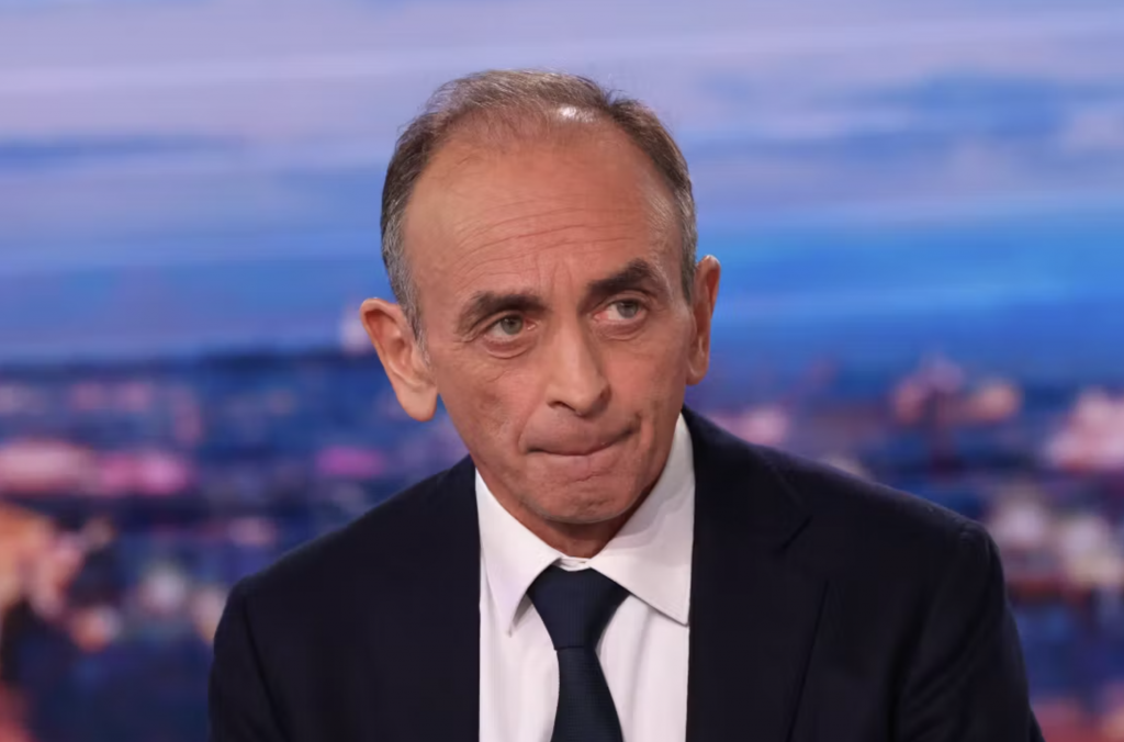 Portrait en gros plan d’un homme politique en costume sombre et cravate, assis sur un plateau de télévision, regard sérieux légèrement tourné sur le côté, avec un décor flou de studio en arrière-plan aux tons bleus et violets.
