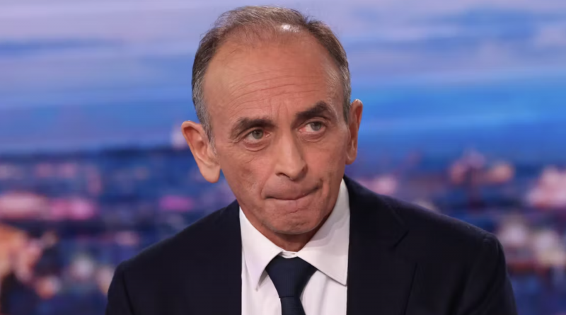 Portrait en gros plan d’un homme politique en costume sombre et cravate, assis sur un plateau de télévision, regard sérieux légèrement tourné sur le côté, avec un décor flou de studio en arrière-plan aux tons bleus et violets.