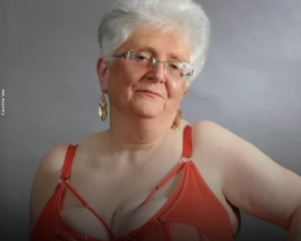 Femme âgée aux cheveux gris courts portant de la lingerie rouge, regard calme et assumé face à l’objectif, posture droite, expression sereine, fond neutre