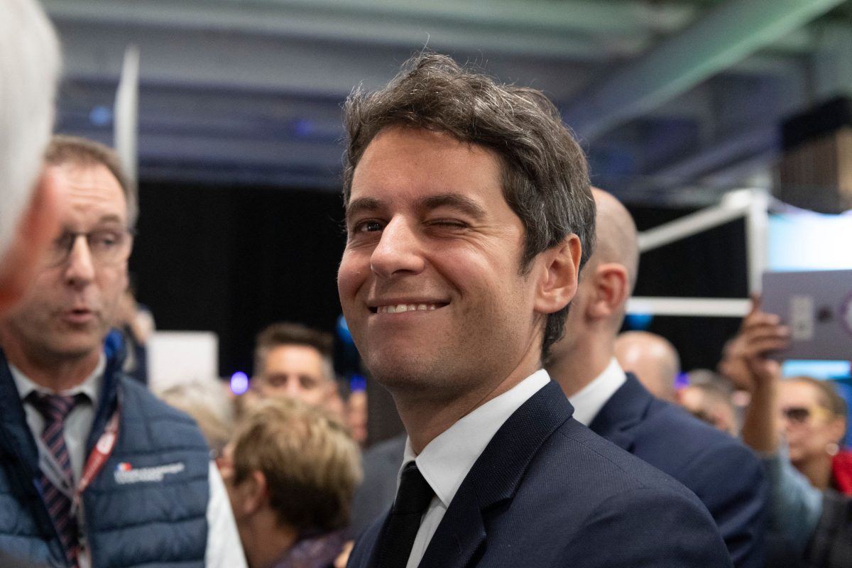 Homme politique en costume sombre et cravate noire, souriant et clignant de l’œil, entouré de plusieurs personnes lors d’un événement public en intérieur.