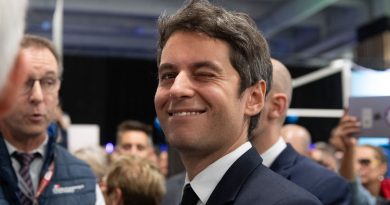 Homme politique en costume sombre et cravate noire, souriant et clignant de l’œil, entouré de plusieurs personnes lors d’un événement public en intérieur.