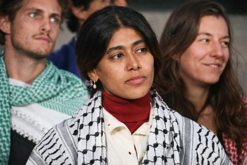 Une élue assise au premier plan lors d’un rassemblement politique, portant un foulard noir et blanc, avec une expression sérieuse, entourée d’autres participants en arrière-plan.