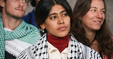 Une élue assise au premier plan lors d’un rassemblement politique, portant un foulard noir et blanc, avec une expression sérieuse, entourée d’autres participants en arrière-plan.