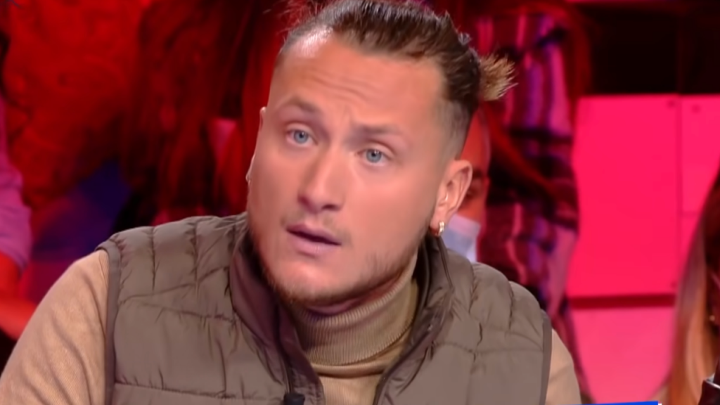 Homme aux cheveux attachés en arrière, portant une veste sans manches matelassée et un pull à col roulé, participant à une émission de télévision sur un plateau éclairé en rouge.