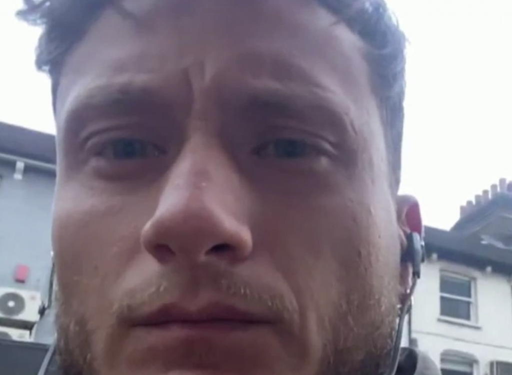 Gros plan sur le visage d’un homme à l’expression grave, filmé en extérieur dans une rue résidentielle, avec des maisons en arrière-plan et un écouteur sans fil visible à l’oreille.