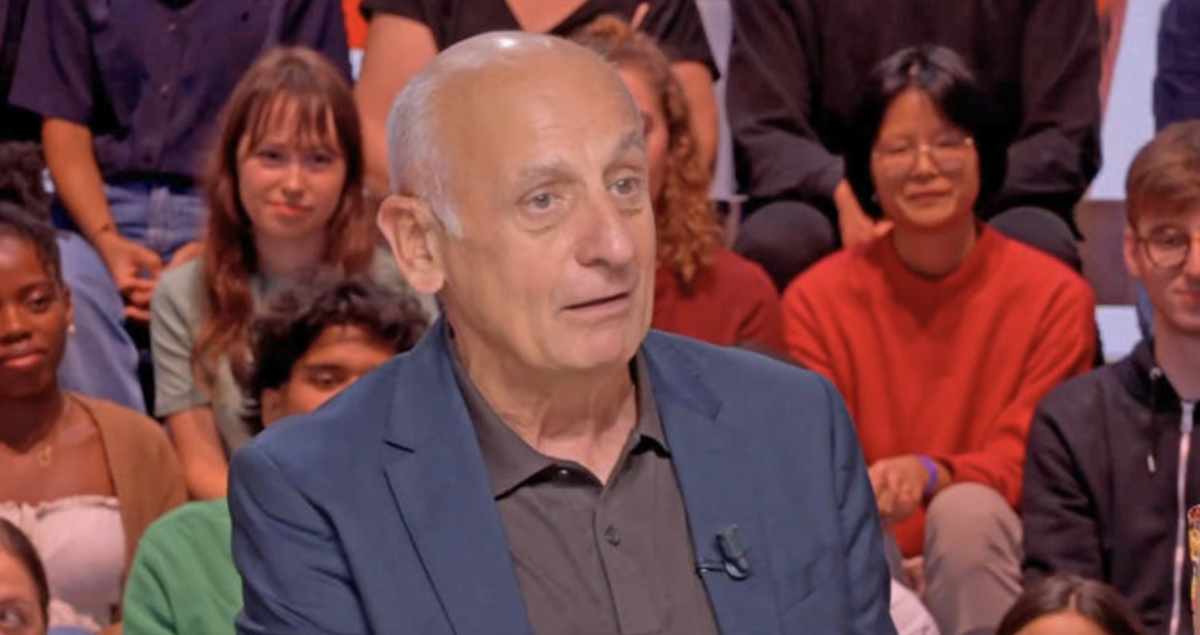Journaliste politique en costume sombre assis sur un plateau de débat télévisé, regard sérieux, avec un public de jeunes adultes en arrière-plan dans un studio lumineux.
