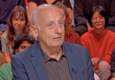 Journaliste politique en costume sombre assis sur un plateau de débat télévisé, regard sérieux, avec un public de jeunes adultes en arrière-plan dans un studio lumineux.
