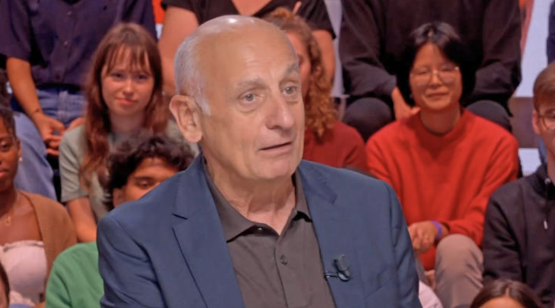 Journaliste politique en costume sombre assis sur un plateau de débat télévisé, regard sérieux, avec un public de jeunes adultes en arrière-plan dans un studio lumineux.