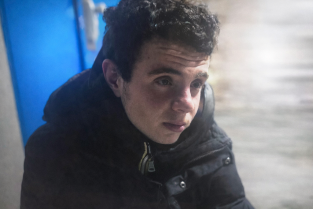 Jeune homme assis à l’extérieur par temps froid, visage marqué par la fatigue, portant un manteau sombre, dans un environnement urbain hivernal.