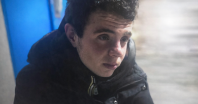 Jeune homme assis à l’extérieur par temps froid, visage marqué par la fatigue, portant un manteau sombre, dans un environnement urbain hivernal.