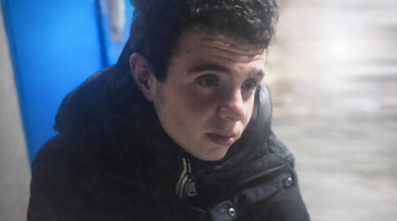 Jeune homme assis à l’extérieur par temps froid, visage marqué par la fatigue, portant un manteau sombre, dans un environnement urbain hivernal.