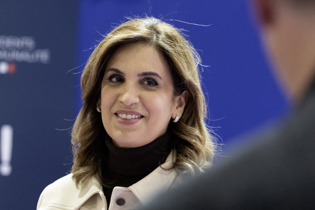 Journaliste souriante lors d’un événement public, tenue claire, fond bleu flouté, éclairage intérieur