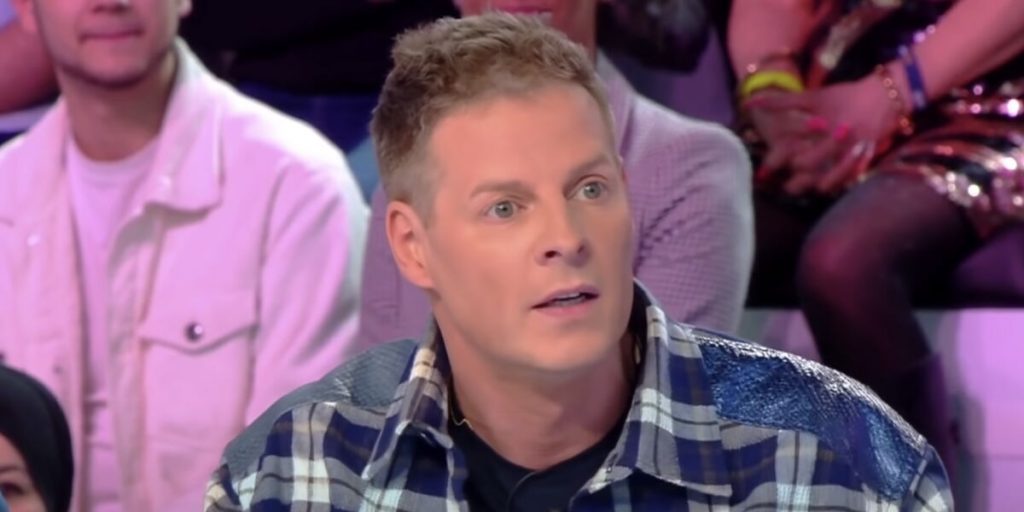 Un homme assis sur un plateau de télévision, regard fixé vers l’avant avec une expression de surprise et de tension, éclairage studio aux tons rosés, public flou en arrière-plan