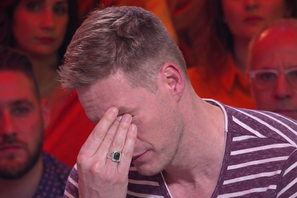 Un homme assis sur un plateau de télévision, la main sur le visage, visiblement bouleversé, sous un éclairage rouge et chaud, avec le public en arrière-plan flouté