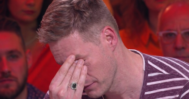 Un homme assis sur un plateau de télévision, la main sur le visage, visiblement bouleversé, sous un éclairage rouge et chaud, avec le public en arrière-plan flouté