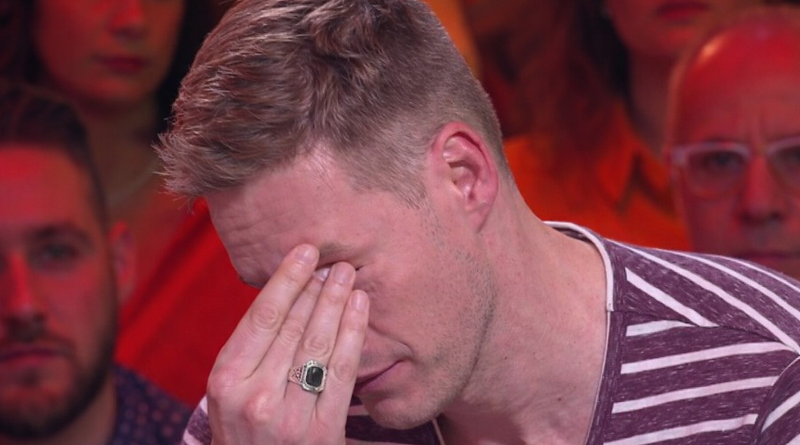 Un homme assis sur un plateau de télévision, la main sur le visage, visiblement bouleversé, sous un éclairage rouge et chaud, avec le public en arrière-plan flouté