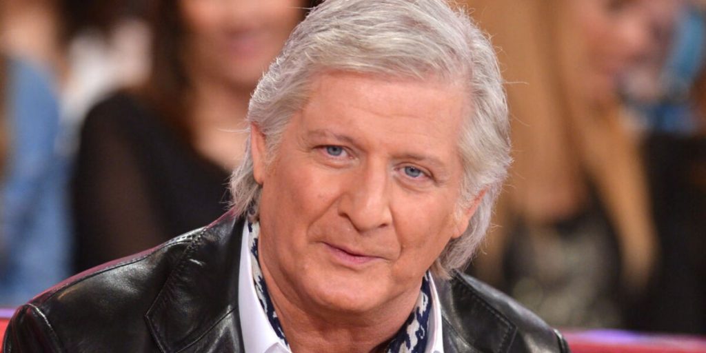 homme aux cheveux gris regardant la caméra, assis sur un plateau de télévision.