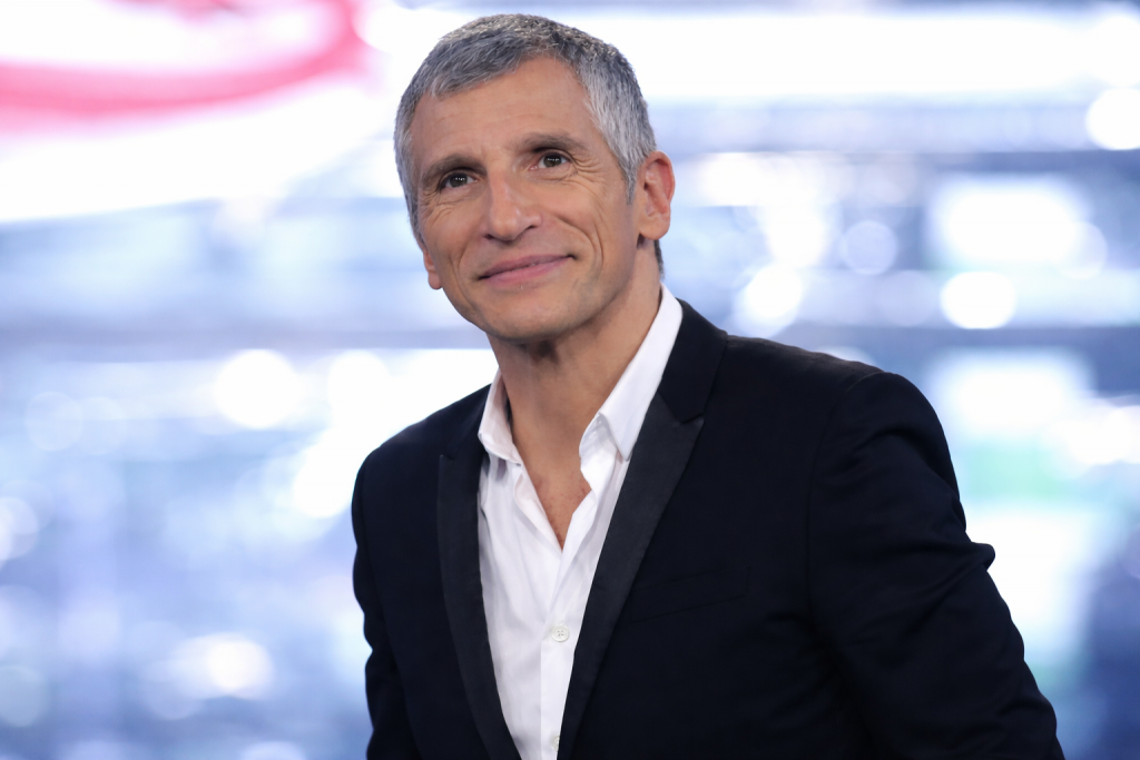 Portrait en plan rapproché d’un animateur de télévision aux cheveux gris courts et au regard expressif, portant un costume noir avec revers satinés et une chemise blanche légèrement ouverte au col, souriant avec assurance sur un plateau de télévision, devant un arrière-plan flou composé de lumières bleues et blanches évoquant un studio professionnel.