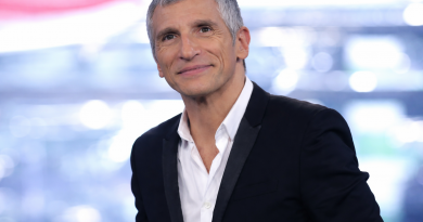 Portrait en plan rapproché d’un animateur de télévision aux cheveux gris courts et au regard expressif, portant un costume noir avec revers satinés et une chemise blanche légèrement ouverte au col, souriant avec assurance sur un plateau de télévision, devant un arrière-plan flou composé de lumières bleues et blanches évoquant un studio professionnel.