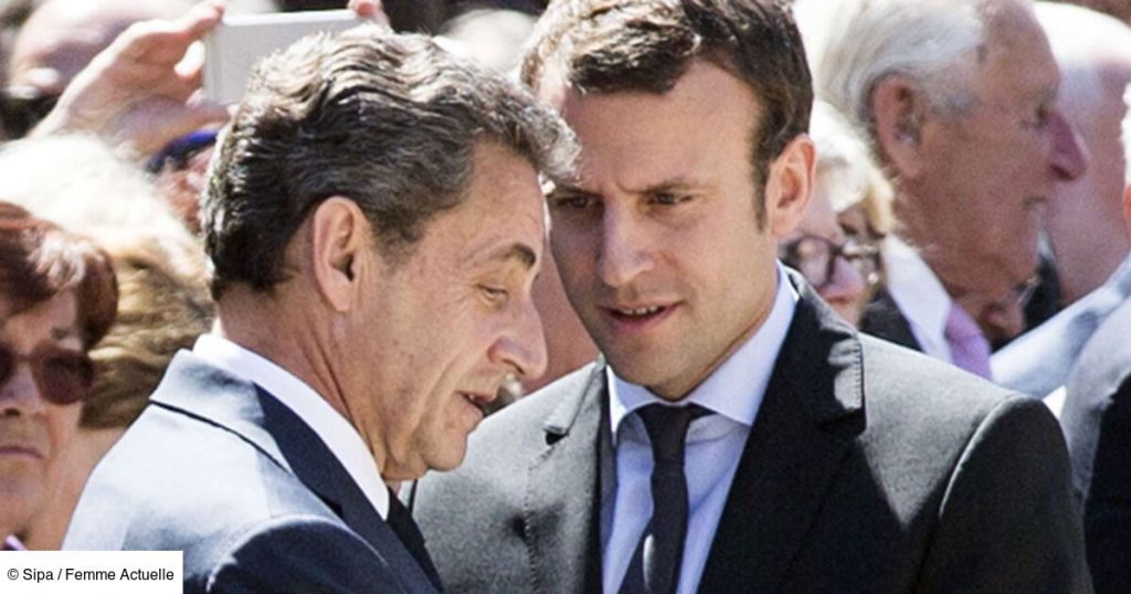 Deux responsables politiques français en costume sombre échangent discrètement lors d’un événement officiel en plein air, entourés d’une foule de participants, avec un arrière-plan flou composé de visages et de smartphones levés.